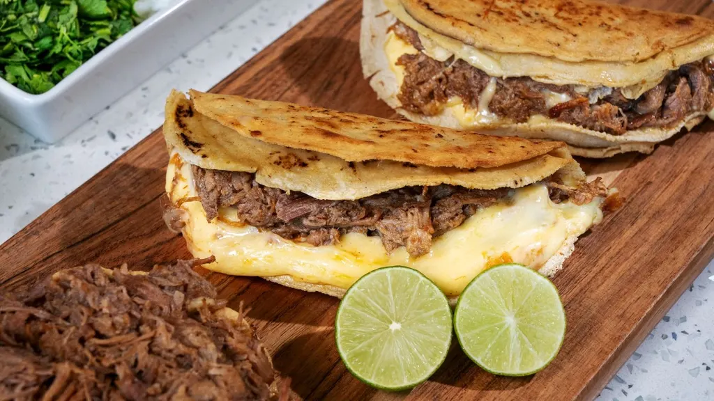 Tacos de birria dorados con queso derretido sobre tabla de madera, con carne deshebrada y limones