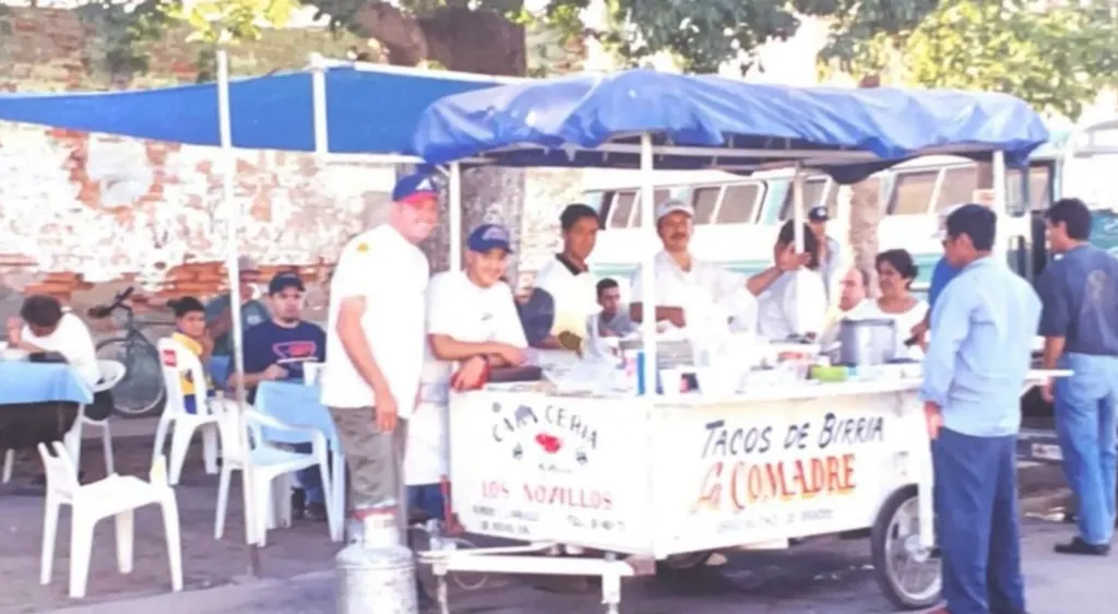 El carrito de Tacos de Birria La Comadre con toldo azul y clientes en la calle, en los años noventa