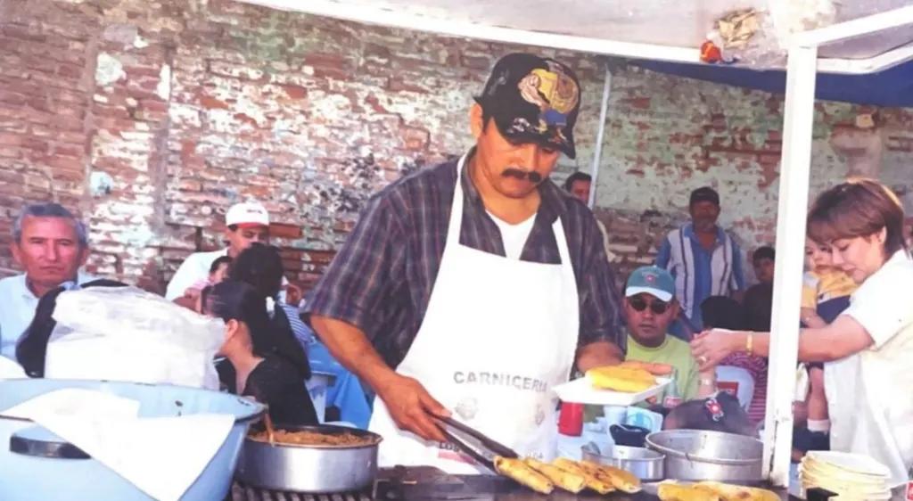 Un taquero con mandil blanco dorando tacos en la plancha bajo el toldo